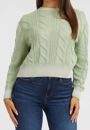 Femme portant un pull torsadé vert clair et un jean bleu foncé, debout avec une main sur le côté, sur un fond uni.