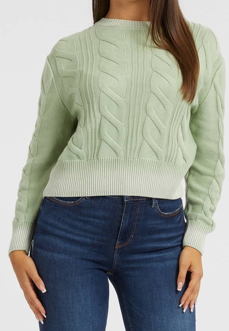 Femme portant un pull torsadé vert clair et un jean bleu foncé, debout avec une main sur le côté, sur un fond uni.