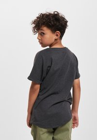 Dreng med krøllet hår iført en mørkegrå t-shirt med korte ærmer og grønne bukser, vendt på siden og ser tilbage over sin skulder på en hvid baggrund.