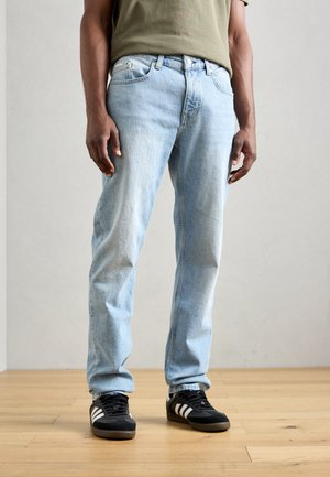 Lichtblauwe denim jeans met een rechte pijp, voorzien van een knoopsluiting, vijf zakken en subtiele vervaging, gedragen met zwarte sportschoenen.