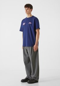 Mörkblå bomullsT-shirt med korta ärmar, med rosa textgrafik. Kombinerad med lösa grå byxor och svarta skor.