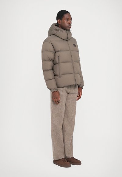 Puffer jacket in taupe, featuring a hood, quilted design, front zipper, and two side pockets. Paired with brown textured trousers and shoes.
