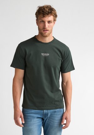 Homme aux cheveux bouclés portant un t-shirt vert foncé Petrol Industries et un jean bleu, debout devant un fond clair uni.