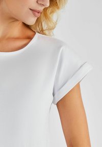 Biała bluzka z krótkim rękawem o okrągłym dekolcie i podwiniętych mankietach, wykonana z gładkiego, elastycznego materiału. Szwy biegnące wzdłuż ramion.
