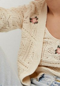 Sweter kardigan w kremowym kolorze z otwartym przodem, charakteryzujący się teksturowanym wzorem i akcentem haftu kwiatowego. Noszony na pasującej bluzce bez rękawów.