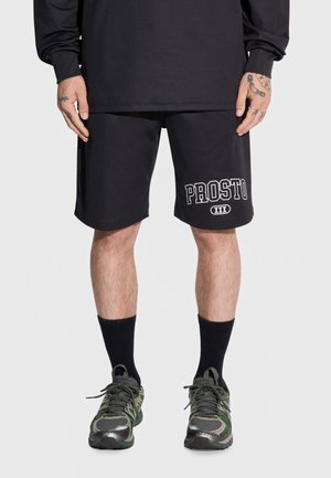 Osoba ubrana w czarne spodenki z napisem „PROSTO XXX”, czarną koszulkę z długim rękawem, czarne skarpetki i zielone buty sportowe moro, stojąca z rękami ułożonymi wzdłuż ciała.