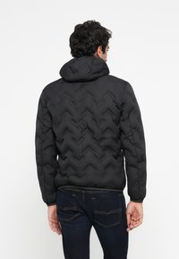 Sort quiltet pufferjakke med et herringbone mønster, hætte og elastiske manchetter. Stoffet ser blødt og struktureret ud og giver isolering.