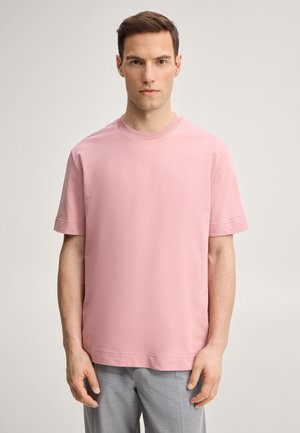 Man standing facing forward, wearing a plain pink short-sleeve T-shirt and light gray pants against a neutral background.