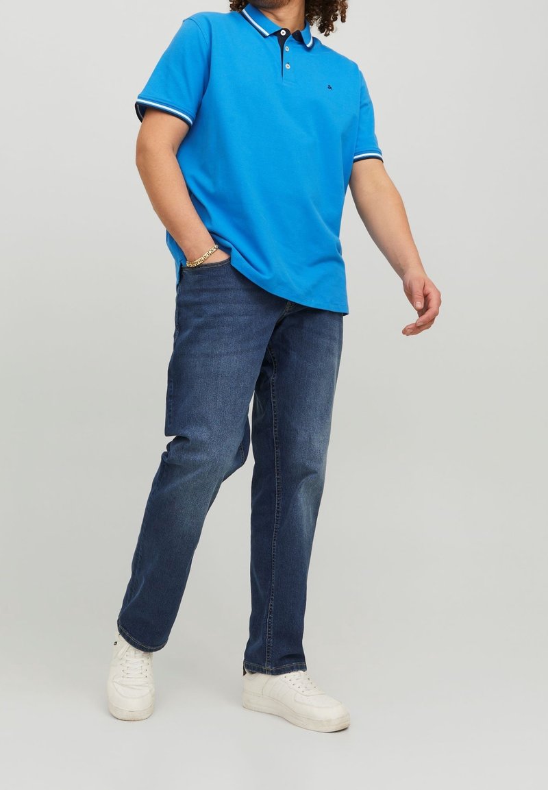 Blå pikétröja med svart krage och vita detaljer, kombinerad med mörka denimjeans och vita sneakers, vilket skapar en avslappnad outfit.