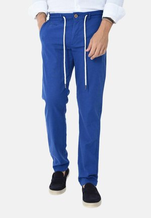 Uomo che indossa pantaloni blu brillanti con coulisse e scarpe nere slip-on, in piedi con una mano in tasca e l’altra rilassata di lato.