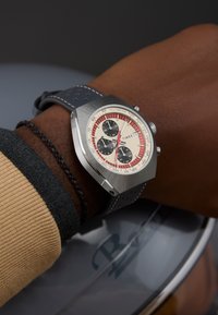 Zilverkleurig horloge van roestvrij staal met een crèmekleurige wijzerplaat, rode tachymeterschaal, zwarte sub-wijzerplaten en een donkerblauwe leren band met perforaties.