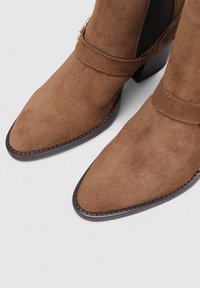 Bottines en suede marron avec des pointes pointues, présentant un détail de sangle et un petit talon carré. La semelle est foncée, apportant contraste et définition.