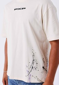 Camiseta de algodón crema con el logo "PXP" en negro, diseño abstracto de salpicaduras de pintura en negro y púrpura cerca del dobladillo, corte relajado, mangas cortas.