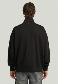 Sweatshirt noir surdimensionné avec un col montant, présentant une petite étiquette logo au dos du cou. Texture lisse et manches côtelées.