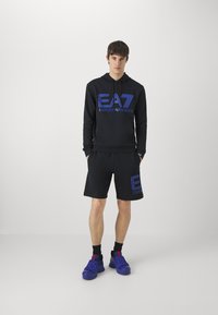 Felpa nera con cappuccio e pantaloni corti con logo EA7 blu, realizzati in tessuto morbido. Abbinati a scarpe sportive blu e calzini neri. Design minimalista.