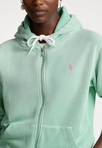 Mentolová zipová mikina s bílým stahovacím šňůrkou, na hrudi s malým růžovým logem a vysoce kvalitní smíšenou bavlněnou látkou.