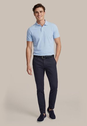 Jonge man glimlacht, draagt een lichtblauw poloshirt, donkerblauwe broek met een zwarte riem en donkerblauwe slip-on schoenen op een neutrale achtergrond.