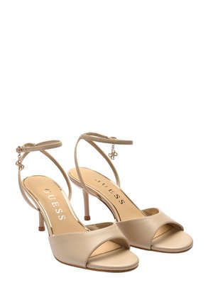 Sandales à talons beiges à bout ouvert avec brides à la cheville et petits charms décoratifs, avec le logo de la marque Guess sur la semelle intérieure.