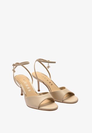 Sandali beige con tacco aperto sulla punta, cinturini alla caviglia e piccoli ciondoli decorativi, con il logo del marchio Guess sulla soletta.