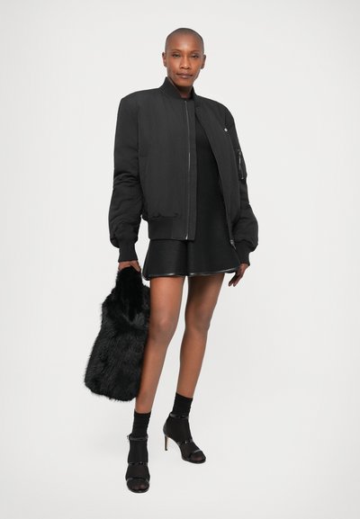 Veste bomber noire avec des poignets côtelés, fermeture éclair et poches latérales, portée sur une robe noire. Sac à main en fourrure noire et chaussures à talons.