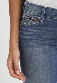 Vaqueros de denim en azul medio, con una textura desgastada, diseño clásico de cinco bolsillos y un pequeño acento a rayas en el bolsillo frontal.