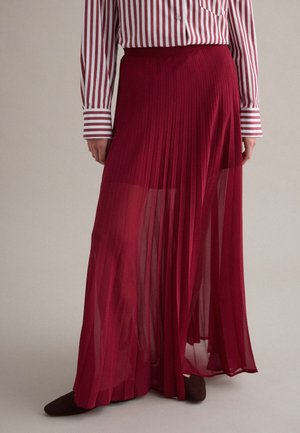 Mujer vestida con una falda larga plisada color rojo intenso translúcido combinada con una camisa de manga larga a rayas verticales rojas y blancas, y zapatos oscuros.