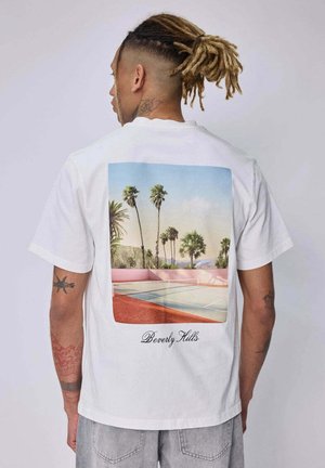 Homme avec des dreadlocks et des tatouages, portant une chemise blanche, montrant un court de tennis coloré à Beverly Hills avec des palmiers en arrière-plan.