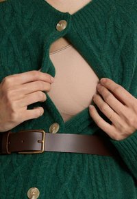 Groene gebreide cardigan met een textuurpatroon, voorzien van een knoopsluiting en een bruine riem. Gelaagd over een naadloze tanktop.