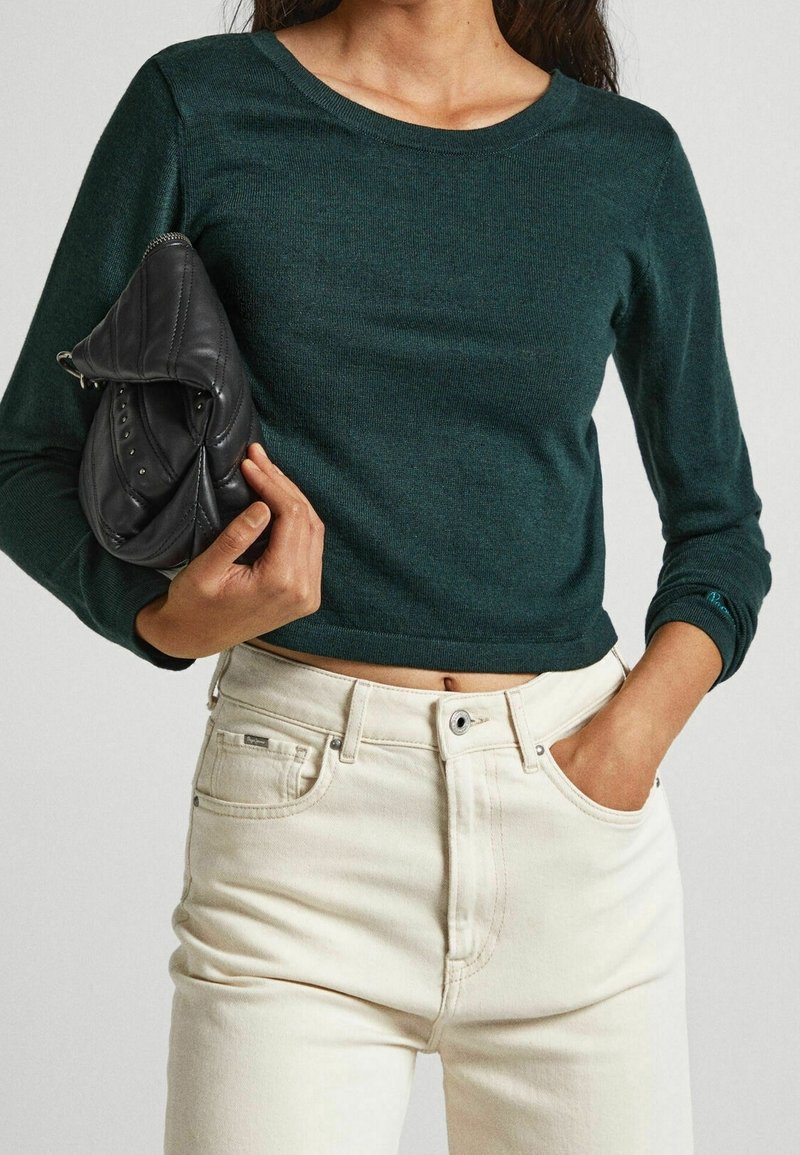 Ciemnozielony, dopasowany sweter z długim rękawem i okrągłym dekoltem, zestawiony z kremowymi, wysokim stanem jeansami. Trzyma czarną skórzaną torbę.