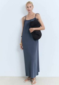 Rochie maxi gri, fără mâneci, cu o croială lejeră, având bretele ajustabile. Modelul poartă o geantă de mână din fibră naturală, de culoare neagră; sandale plata.