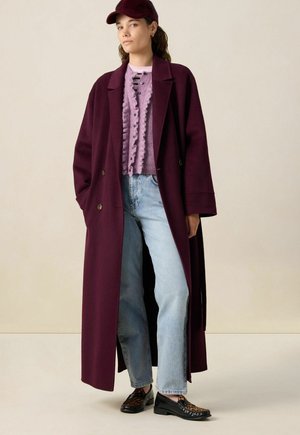 Vrouw in een lange bordeauxrode jas, paarse gerimpelde cardigan, lichtblauwe jeans, loafers met luipaardprint en een bijpassende bordeauxrode baret op een neutrale achtergrond.