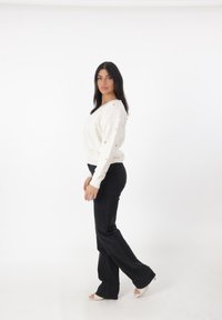 Maglione bianco lavorato a maglia con motivo a cuore dorato testurizzato, abbinato a jeans neri svasati e sandali con tacco trasparente. Sfondo semplice.