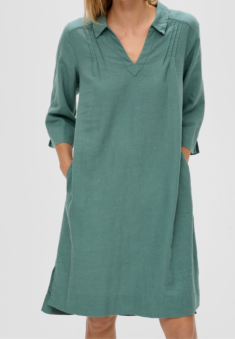 Femme portant une robe verte ample, longueur genou, avec des manches trois-quarts et un col en V, les mains dans les poches latérales.