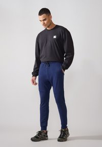 Svart langermet skjorte med en liten logo, kombinert med marineblå joggebukser med snøring i midjen og teksturerte aksenter, fullført med svarte joggesko.