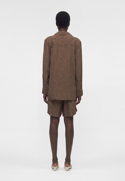 Άτομο που στέκεται με την πλάτη προς τα εμπρός, φορά καφέ μπλούζα μακρυμάνικη και ταίριαζο σορτς με αφηρημένα γραμμικά σχέδια και μπεζ μυτερά τακούνια.