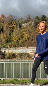 Maglietta sportiva a maniche lunghe blu con dettagli bianchi, abbinata a leggings neri con strisce riflettenti. In piedi accanto a un fiume con fogliame autunnale.