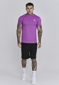 Paarse, aansluitende sporthemd met korte mouwen, gecombineerd met zwarte shorts. Witte sneakers en zichtbare tatoeages op armen en benen.