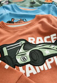Sweat-shirt orange avec un motif texturé de voiture de course en vert, comprenant des étoiles blanches et le texte en gras "CHAMPION DE COURSE". Superposé avec des vêtements bleus et verts.