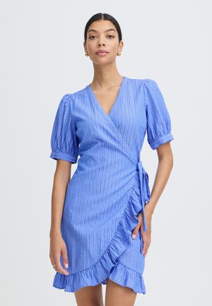 Kobieta w niebieskiej, krótkorękawowej sukience kopertowej z pionowymi paskami i falbanką na dole, na jednolitym tle.