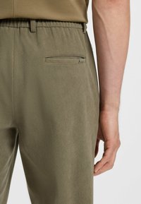 Olijfgroene broek met een geribbelde textuur, voorzien van een sleutelzakje op de achterkant en een rechte pasvorm. De tailleband heeft verstelbare details.