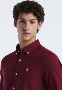 Bordeaux knoopshirt van glad katoen, met een klein wit pinguïnlogo op de linkerborst en contrasterende knopen op de kraag.