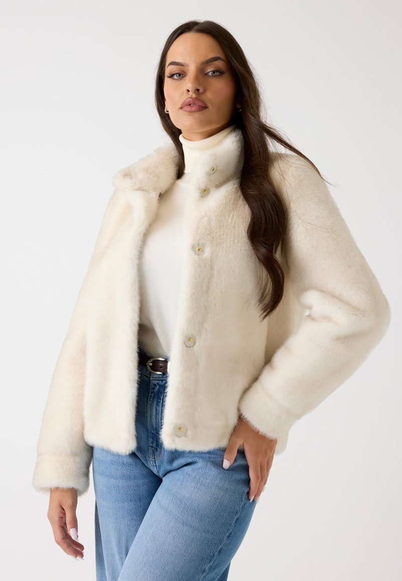 Cremfarbene Kunstfelljacke mit weicher Textur, mit Kragen, Goldknopfverschluss und kurzen Ärmeln, kombiniert mit hellen Jeans.