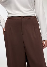 Brązowe spodnie szyte na miarę, wykonane z gładkiego materiału, z zapinaniem na guzik z przodu, bocznymi kieszeniami oraz wyraźnym zagięciem wzdłuż każdej nogawki.