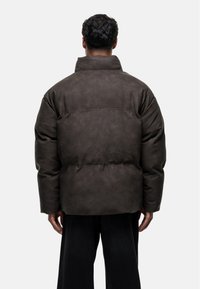 Doudoune oversize marron avec un col haut et une texture lisse. Présente un motif brodé dans le dos et des manches larges.