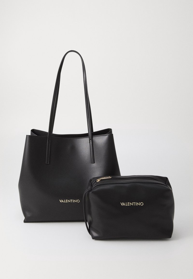 Sac cabas en cuir noir Valentino avec longues anses et pochette noire assortie zippée avec logos dorés sur un fond neutre.