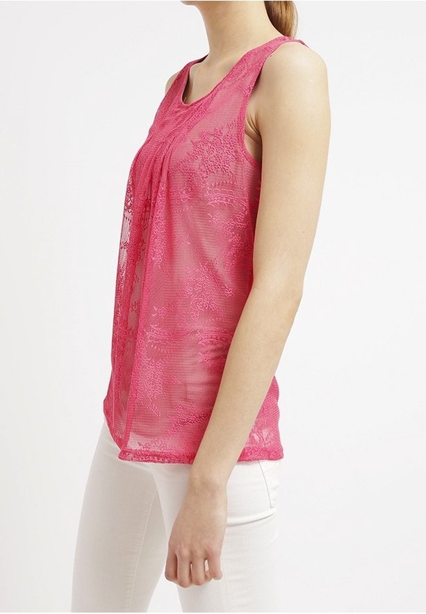 Femme portant un haut en dentelle rose sans manches et un pantalon blanc, debout de profil sur un fond blanc uni.