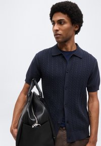 Άνδρας με κοντά σγουρά μαλλιά φορά μπλε ναυτικό πλεκτό πουκάμισο με κουμπιά, κρατά μια μεγάλη μαύρη δερμάτινη τσάντα με ορατή τσέπη με φερμουάρ.