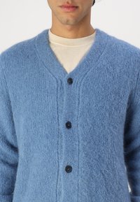 Μπλε κασκόλ φτιαγμένο από απαλό, υφασμάτινο υλικό. Διαθέτει βαθύ V-λαιμό και τέσσερα μαύρα κουμπιά μπροστά. Ραφές με ραβδώσεις στα μανίκια.