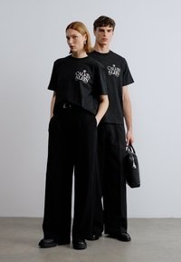 Μαύρα T-shirt με λευκό λογότυπο, συνδυασμένα με φαρδιά μαύρη παντελόνια. Το ανδρικό μοντέλο κρατά μια μικρή μαύρη τσάντα. Μινιμαλιστικός σχεδιασμός και χρωματική παλέτα.