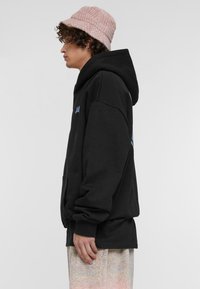 Sudadera con capucha negra y bolsillo frontal. Material suave, corte holgado y logo azul en contraste en el pecho. Lleva puesta una gorra tipo cubo de canalé rosa.